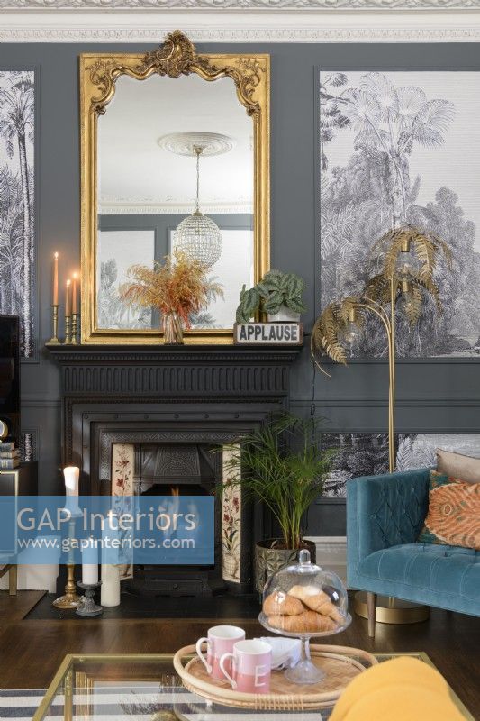 Black painted Victorian fireplace with gold mantle mirror in a grey painted living room with paneling filled with monochrome tropical tree wallpaper
